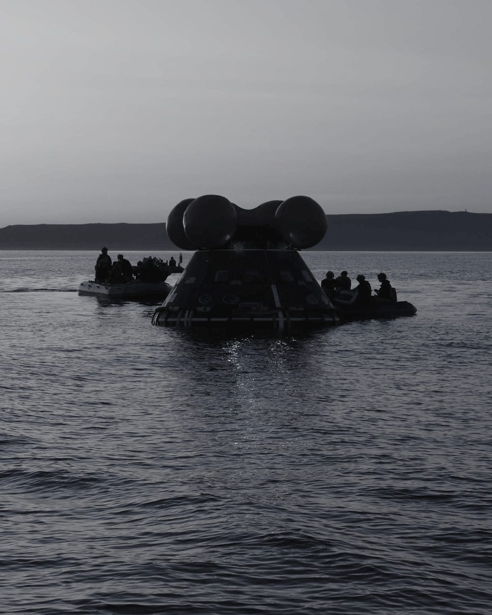 Recovery crews in small boats beside a floating space capsule at dusk